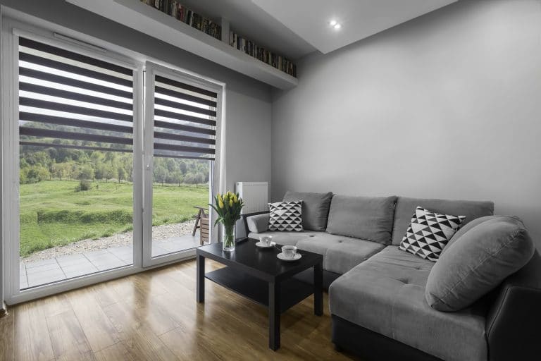 Modernes Wohnzimmer mit grauem Sofa, Couchtisch, Zubehör und großen Fenstern mit Blick ins Grüne.