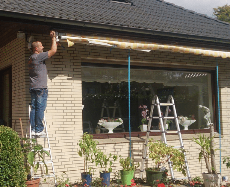 Mann auf einer Leiter, der im Rahmen eines Notdienstes eine Markise über einem Fenster eines Wohnhauses installiert.