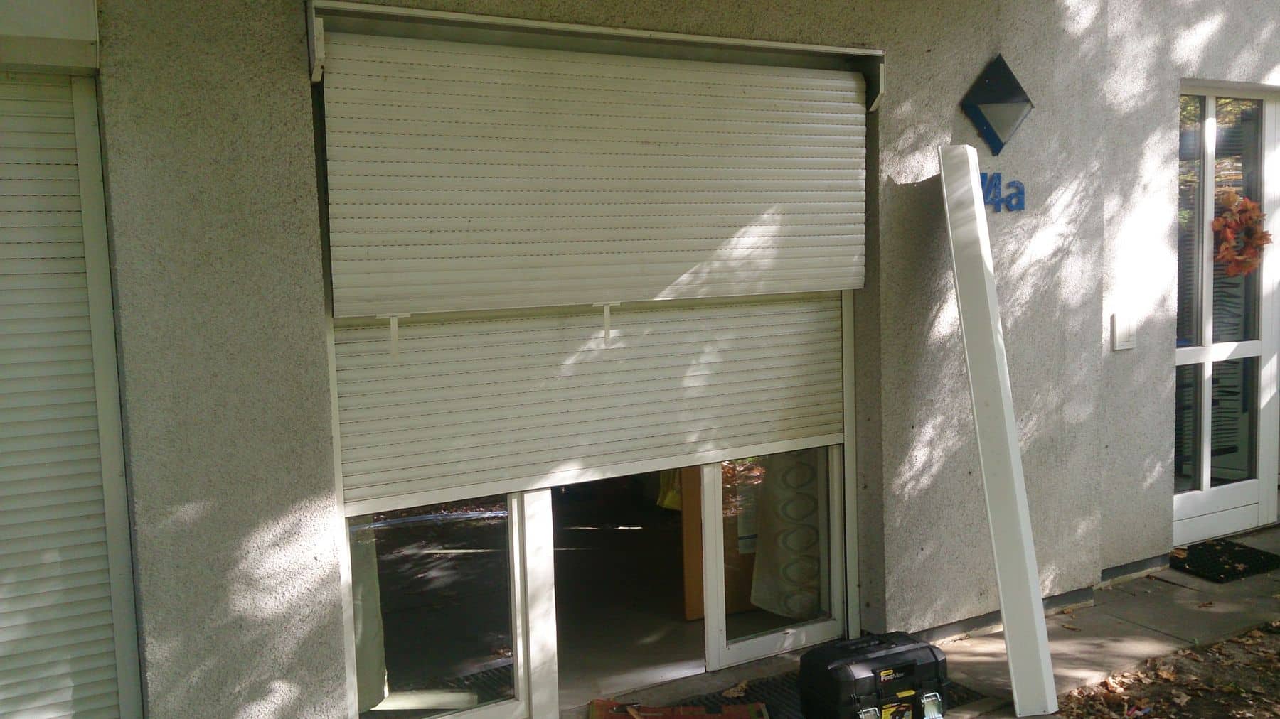 Ein Fenster mit halb geschlossenen Jalousien, die einen Schatten auf die Außenwand werfen, daneben eine offene Tür mit verschiedenen Gegenständen, darunter eine Leiter, auf dem Boden in der Nähe.
