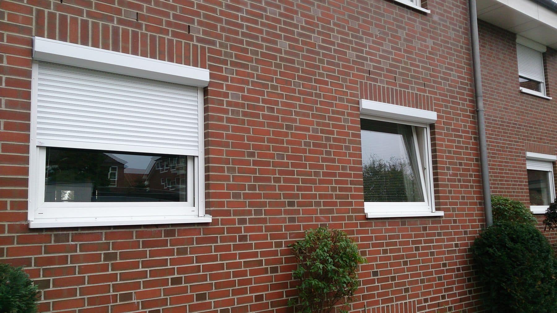 Zwei Fenster mit weißen Rahmen an einer roten Backsteinmauer, eines mit geschlossenen Jalousien und das andere offen zur Sicht.