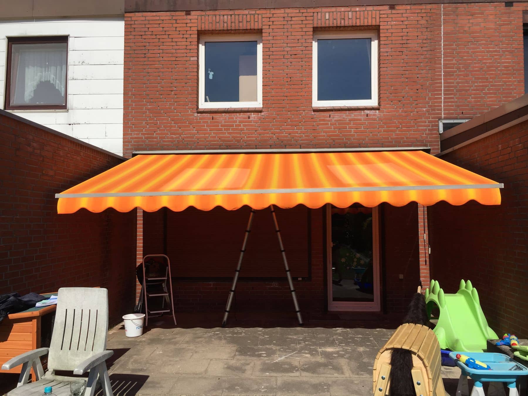 Leuchtend orangefarbene Markise über der Terrasse eines Backsteinhauses, mit einer Leiter und verschiedenen verstreuten Gegenständen.